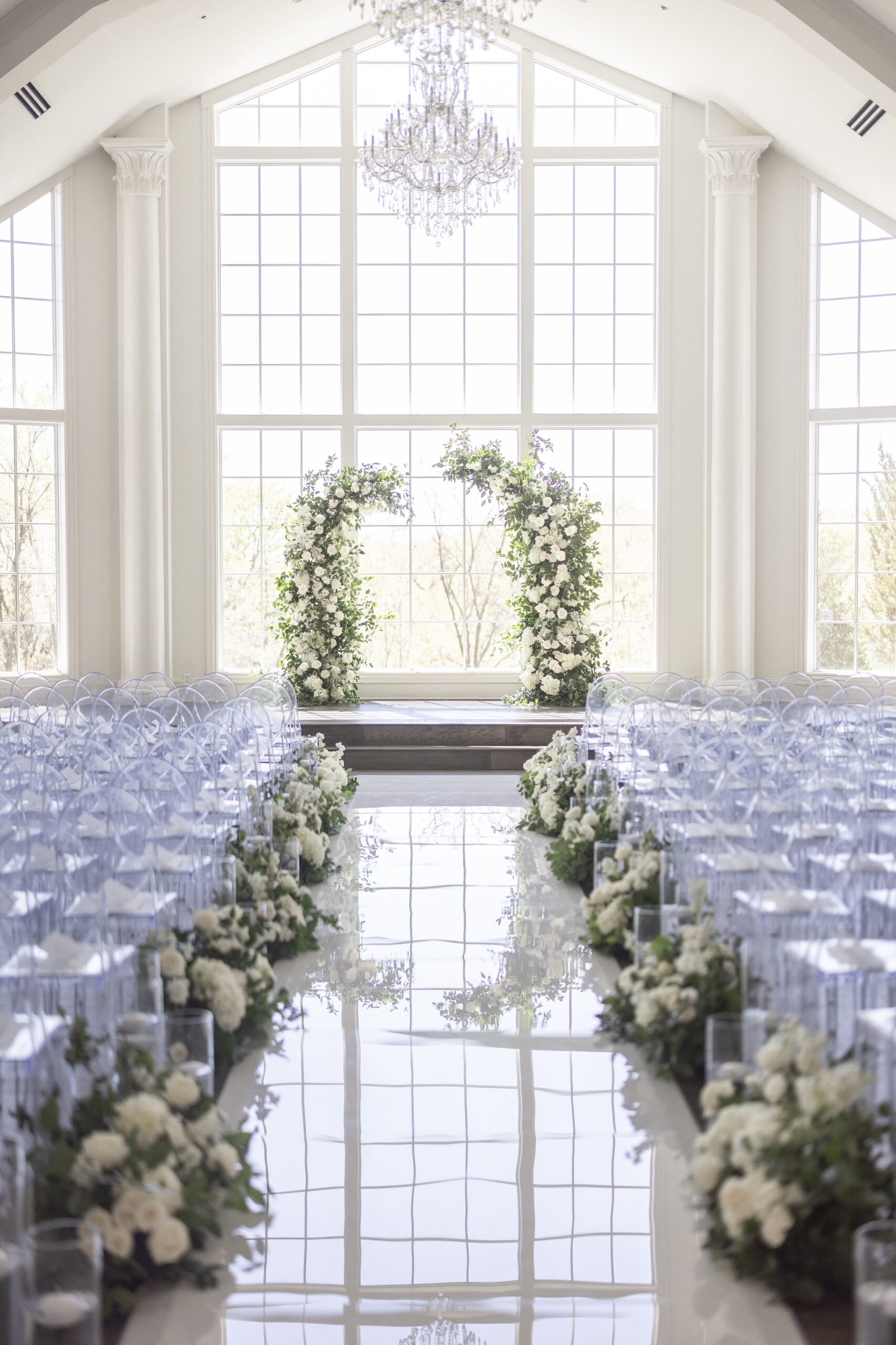 Ceremony spot with floor to ceiling windows, acrylic chairs, a floral arch, and a floral-lined aisle.