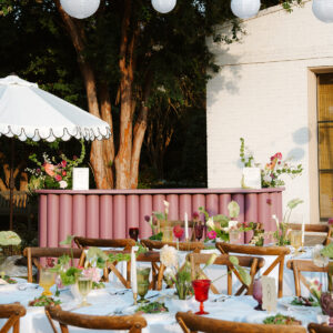 A pink ribbed bar facade, white umbrella, hanging paper lanterns, and white linen tables with wooden chairs.