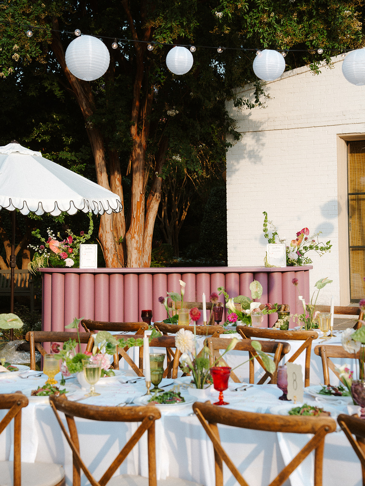 A pink ribbed bar facade, white umbrella, hanging paper lanterns, and white linen tables with wooden chairs.