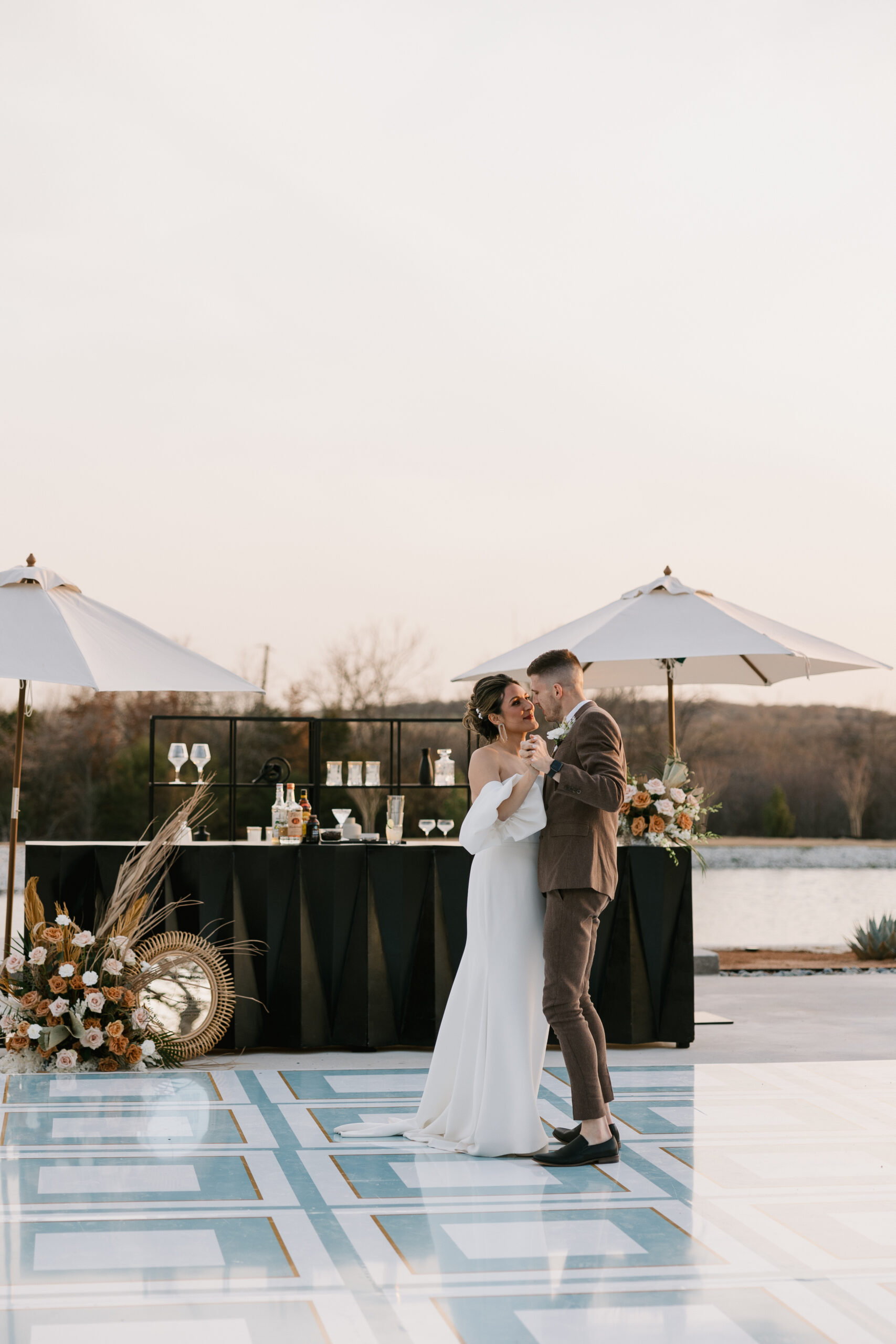 A bride and groom share a first dance outside during the sunset in front of a custom bar and florals.