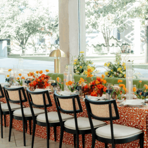 Black and rattan chairs pulled up to an orange printed linen table with bright orange and yellow florals and gold lamps.
